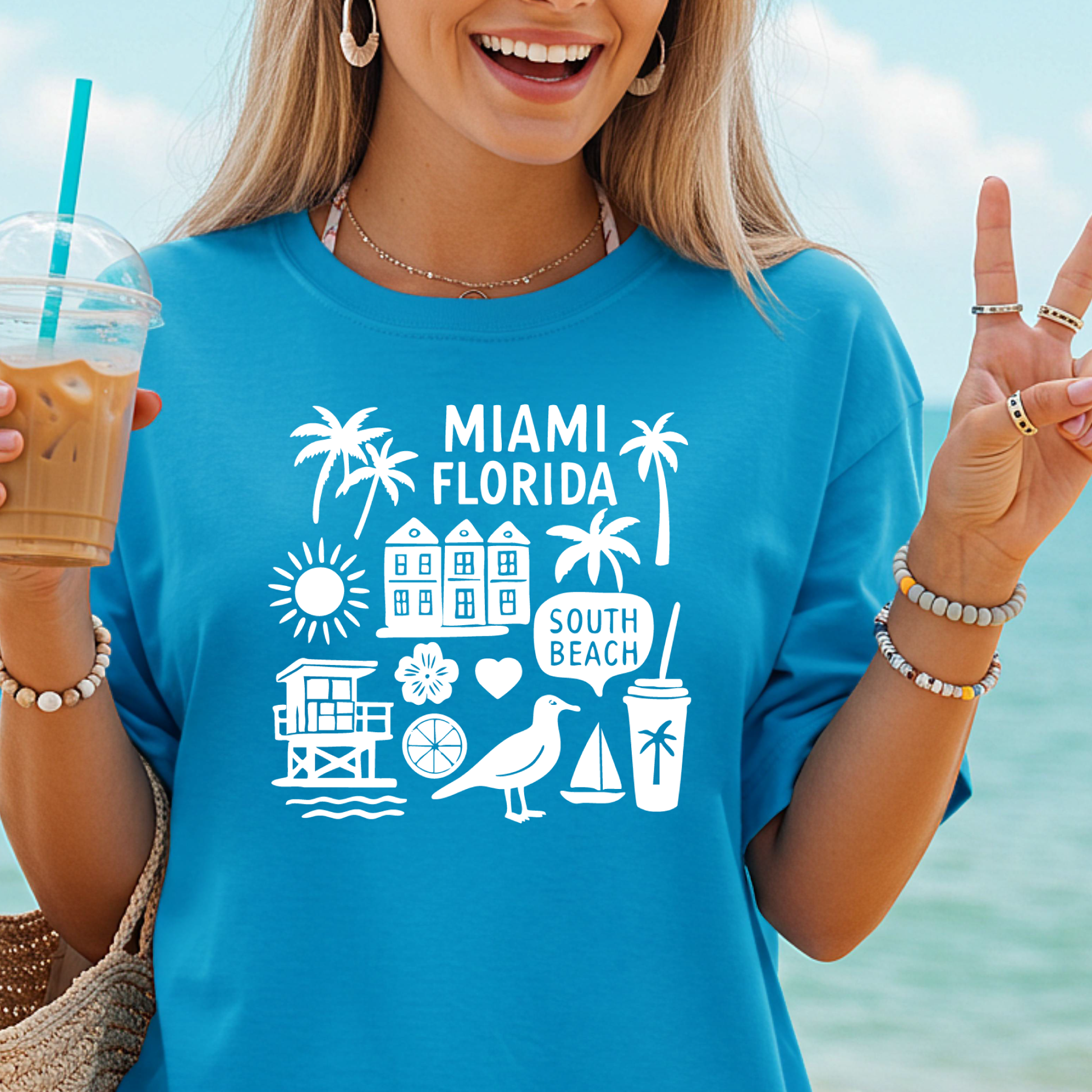 Woman wearing a blue 'Miami Florida' t-shirt with beach-themed graphics, holding a drink by the ocean.