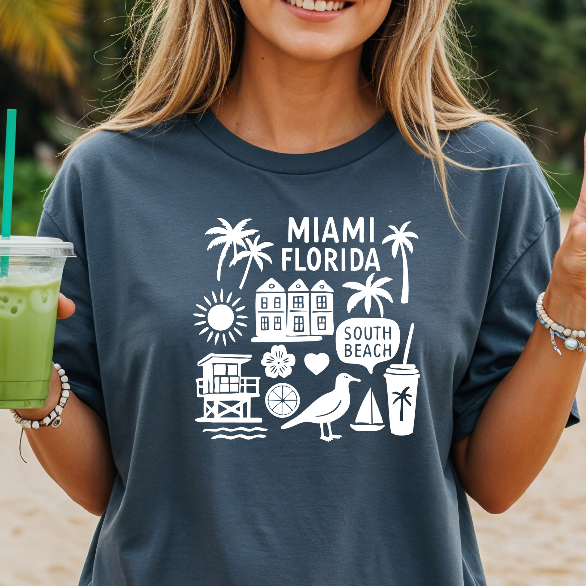 Person wearing a navy blue t-shirt with 'Miami Florida' and beach icons, holding a green drink.