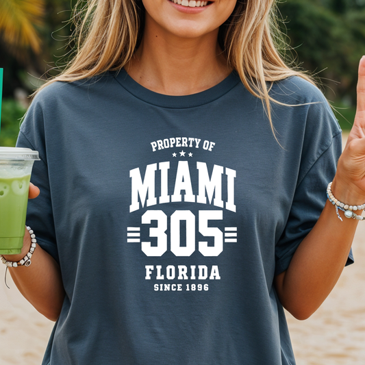 Person wearing a navy blue t-shirt with 'Property of Miami 305 Florida Since 1896' text, holding a green drink.