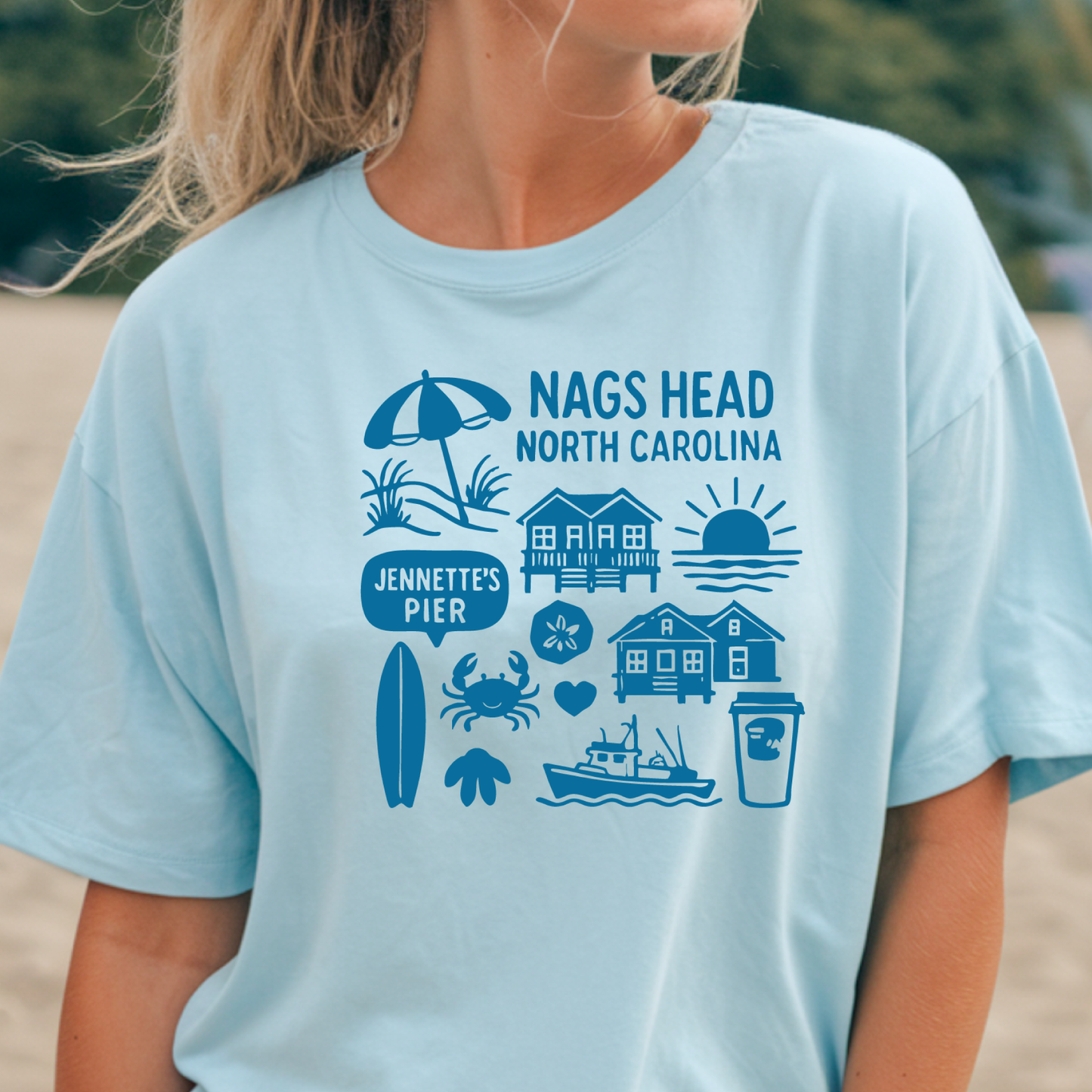 Light blue t-shirt with Nags Head, North Carolina, and Jennette's Pier graphics on a blurred beach background.