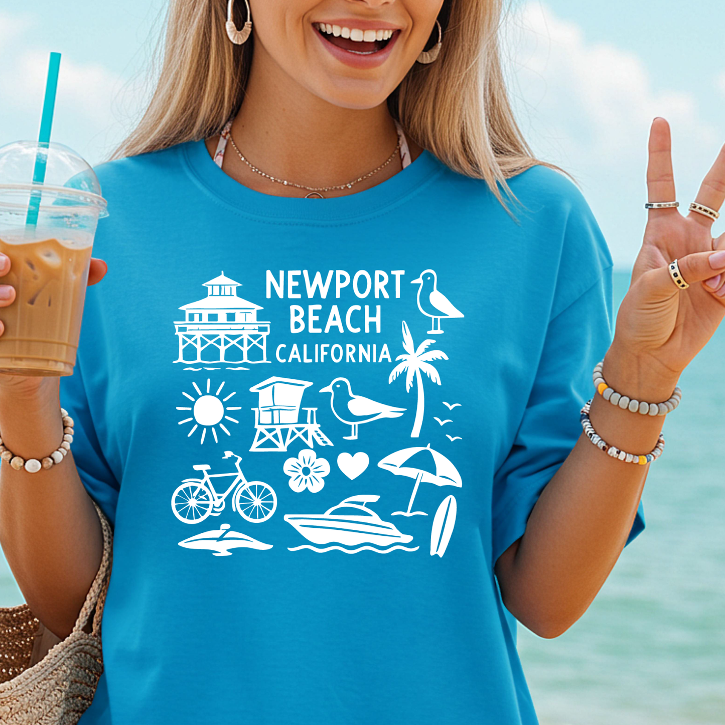 Woman wearing a blue t-shirt with 'Newport Beach California' design, holding a drink by the beach.