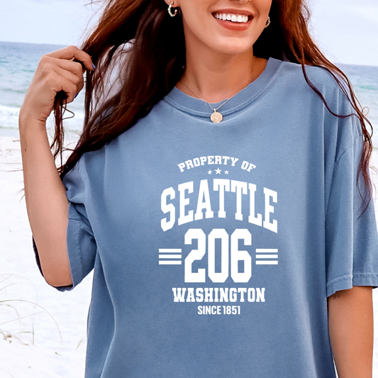Woman wearing a blue t-shirt with 'Property of Seattle 206 Washington Since 1851' text on a beach.
