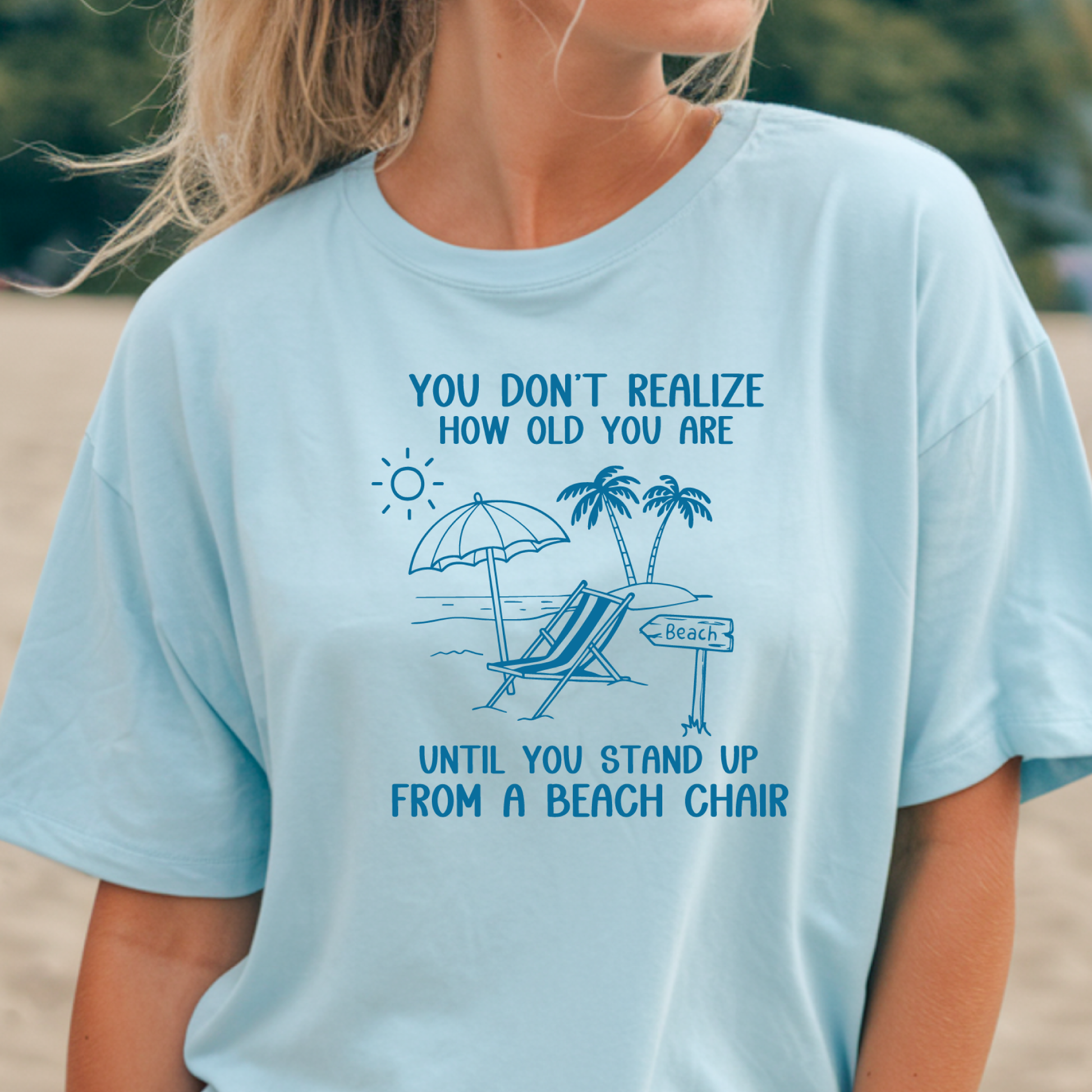 Person wearing a light blue t-shirt with a beach-themed graphic and text on a blurred beach background