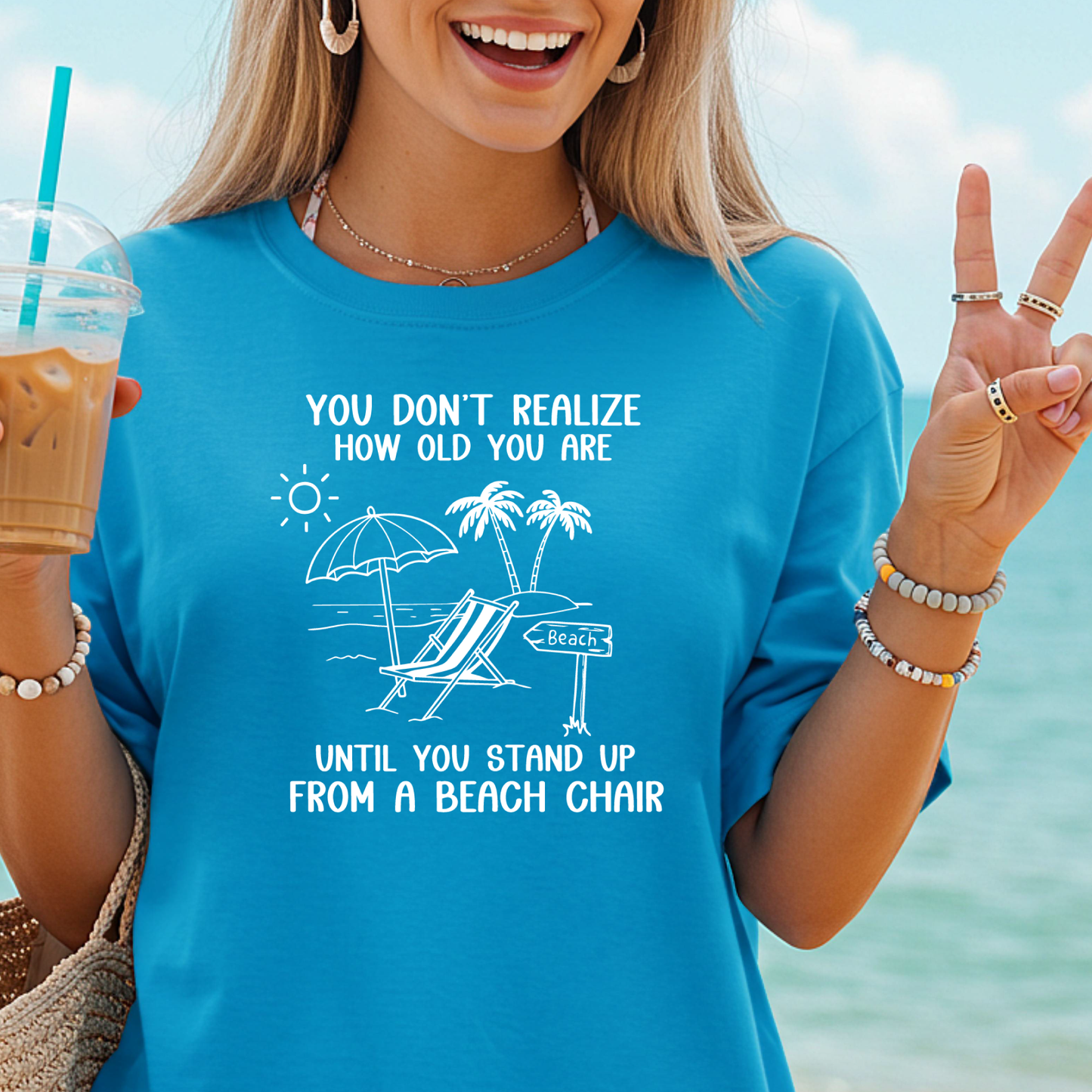 Person wearing a blue t-shirt with a beach-themed design and text, standing by the ocean.