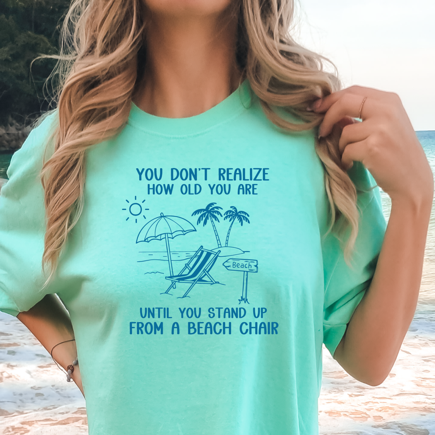 Person wearing a light green t-shirt with a beach-themed design and text at a beach.