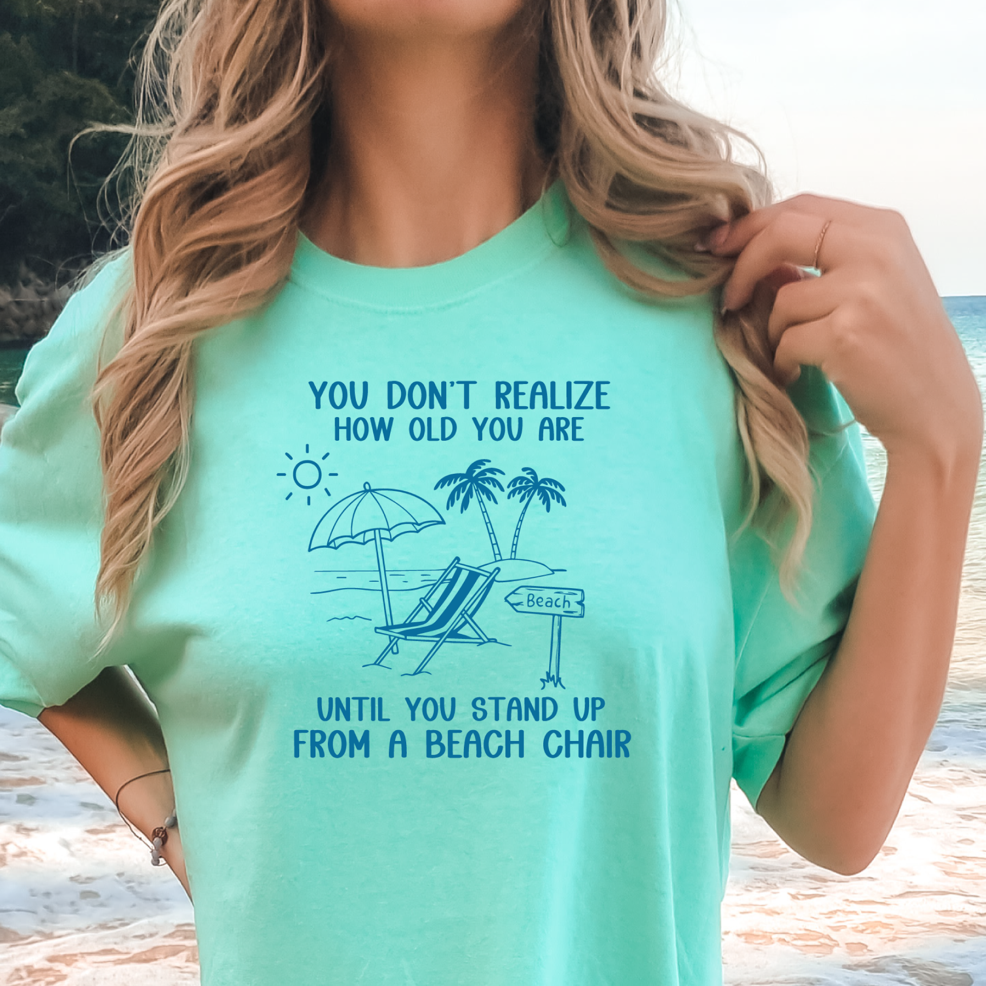 Person wearing a light green t-shirt with a beach-themed design and text at a beach.