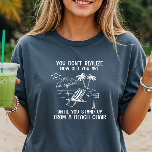 Person wearing a navy blue t-shirt with a beach-themed graphic and text, holding a drink on a beach.
