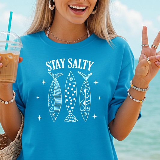 Person wearing a blue t-shirt with 'Stay Salty' and fish graphics, holding a drink by the beach.