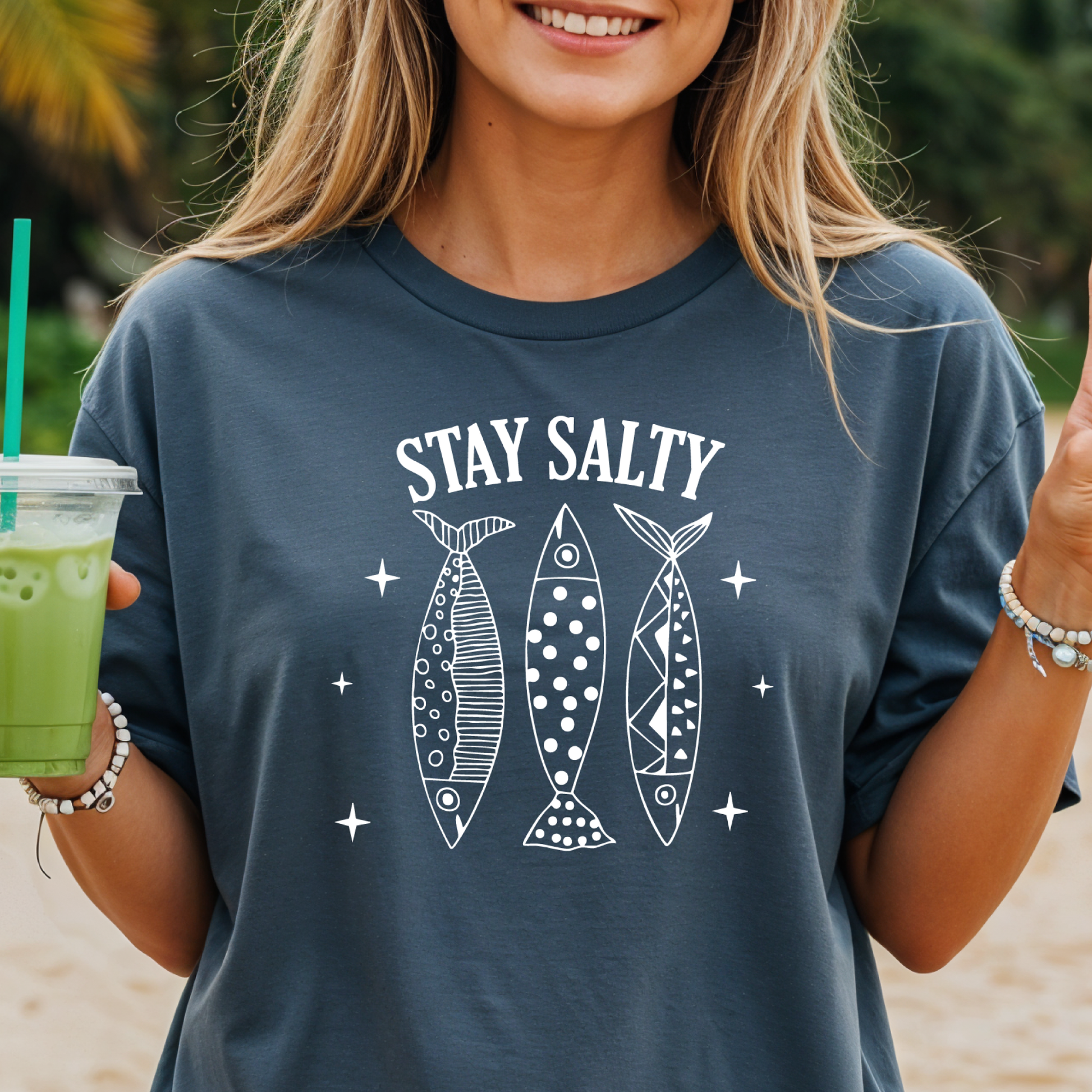 Person wearing a navy blue t-shirt with 'Stay Salty' and fish illustrations, holding a green drink.