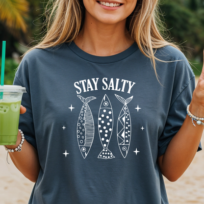 Person wearing a navy blue t-shirt with 'Stay Salty' and fish illustrations, holding a green drink.