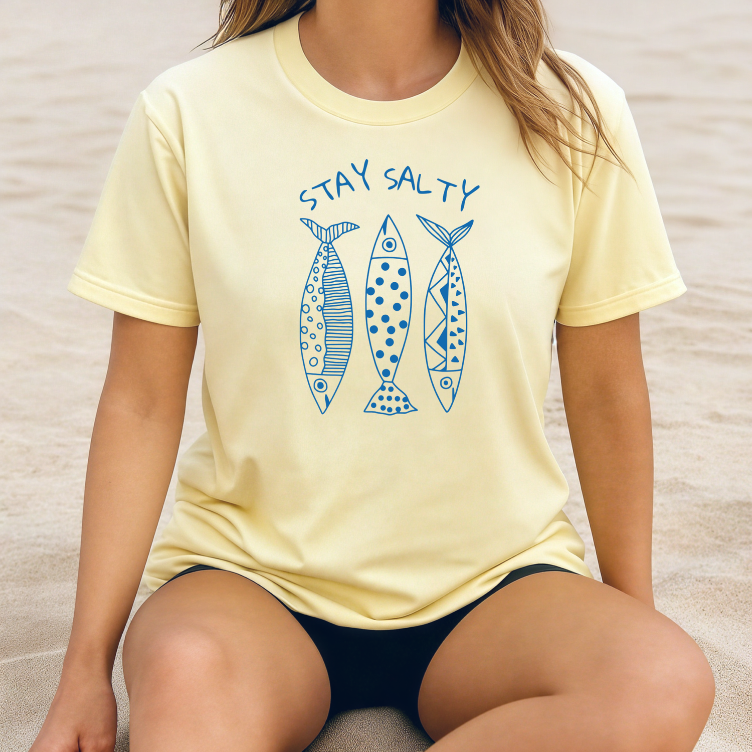Comfort Colors 1717 "Stay Salty" t shirt with three artsy designed sardines one color graphic. Picture of a girl sitting on the beach wearing an oversized t shirt.
