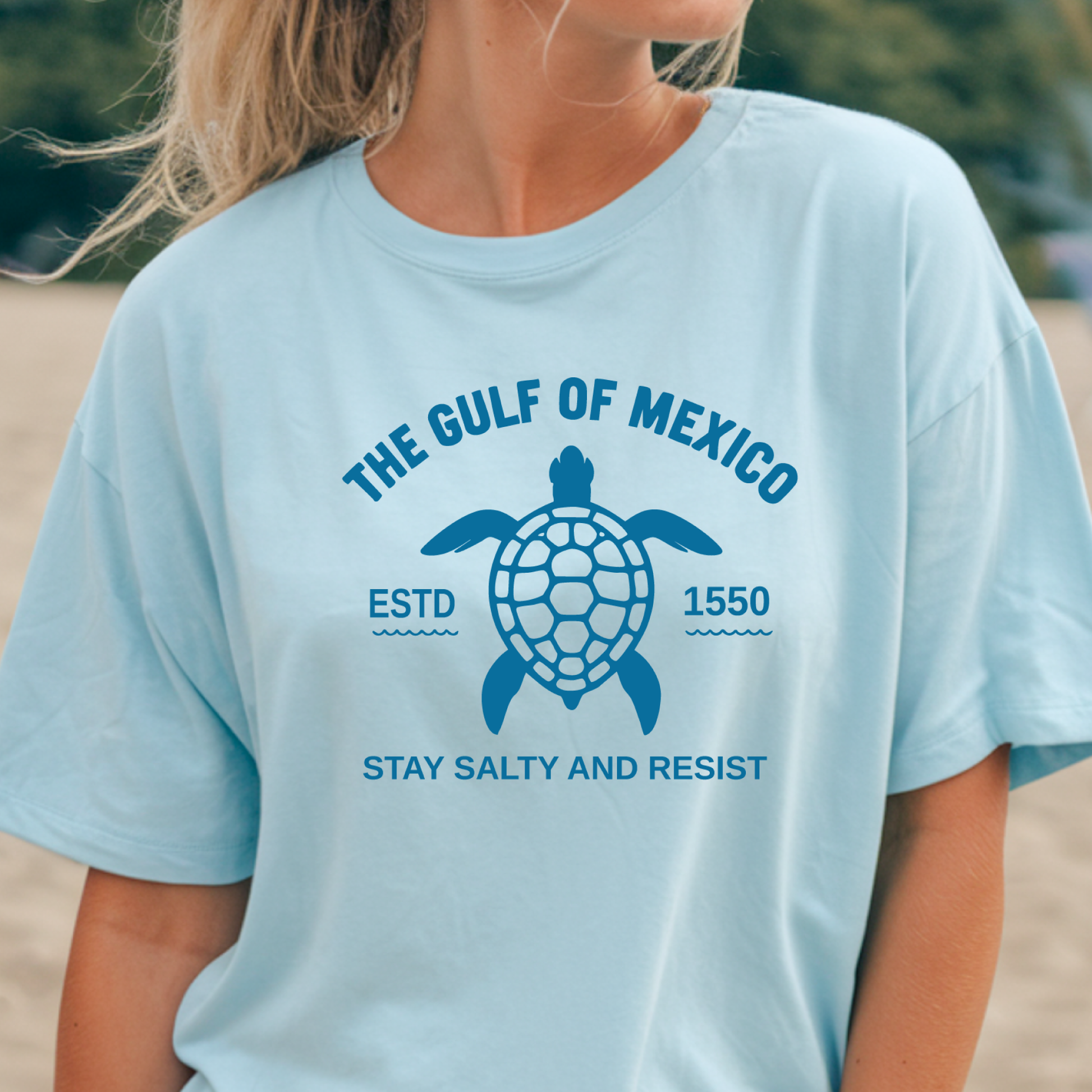 Light blue t-shirt with turtle graphic and text on a blurred beach background