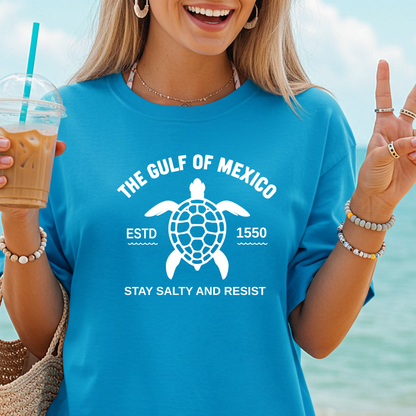 Person wearing a blue t-shirt with a turtle graphic and text by the beach