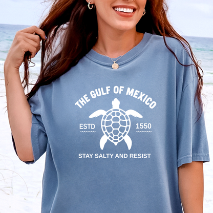 Person wearing a blue t-shirt with a turtle graphic and text on a beach.