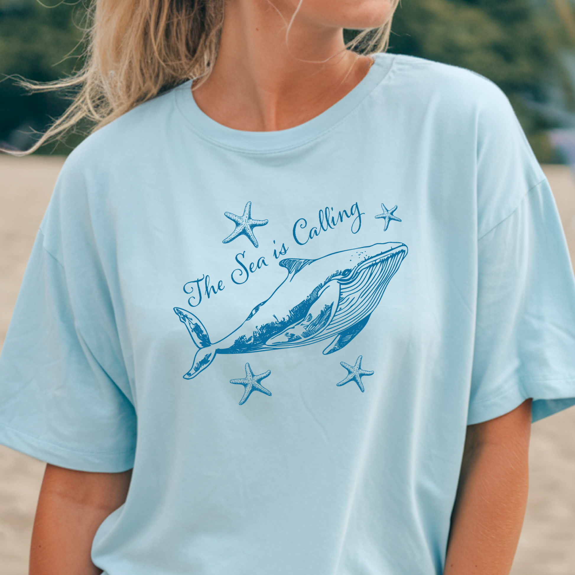 Comfort Colors t-shirt featuring a blue whale illustration surrounded by starfish and the handwritten-style text “The Sea is Calling.” The detailed marine artwork creates a calming coastal aesthetic. This unisex garment-dyed tee offers a soft vintage feel and relaxed fit, perfect for ocean lovers, beach trips, and anyone who enjoys sea-themed graphic shirts with a coastal vibe.