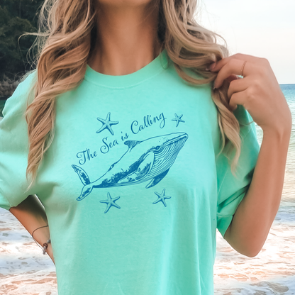 Comfort Colors t-shirt featuring a blue whale illustration surrounded by starfish and the handwritten-style text “The Sea is Calling.” The detailed marine artwork creates a calming coastal aesthetic. This unisex garment-dyed tee offers a soft vintage feel and relaxed fit, perfect for ocean lovers, beach trips, and anyone who enjoys sea-themed graphic shirts with a coastal vibe.