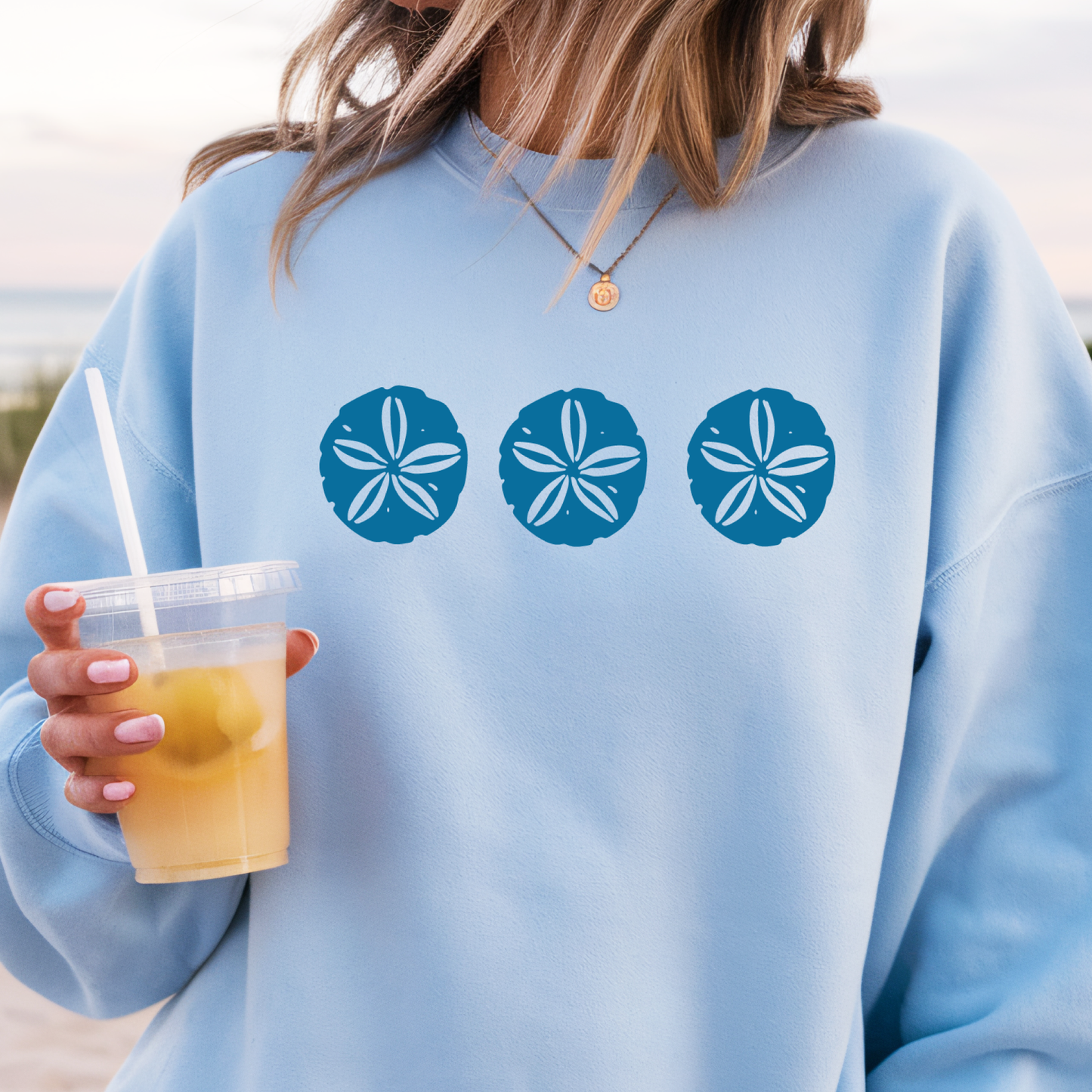 Gildan sweatshirt featuring three blue sand dollar graphics printed across the chest. Soft cotton-poly blend fabric with ribbed cuffs and waistband for a cozy, durable fit. Perfect for beach lovers and coastal style enthusiasts who appreciate simple ocean-inspired designs. Ideal for layering on cool seaside evenings or adding a subtle beach vibe to casual outfits.