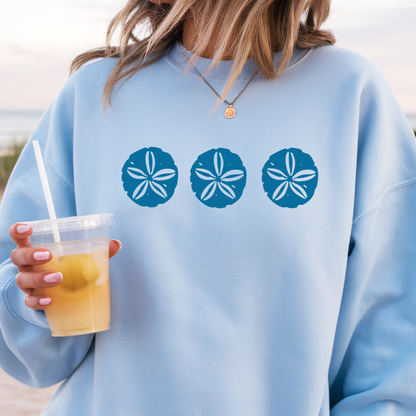 Gildan sweatshirt featuring three blue sand dollar graphics printed across the chest. Soft cotton-poly blend fabric with ribbed cuffs and waistband for a cozy, durable fit. Perfect for beach lovers and coastal style enthusiasts who appreciate simple ocean-inspired designs. Ideal for layering on cool seaside evenings or adding a subtle beach vibe to casual outfits.