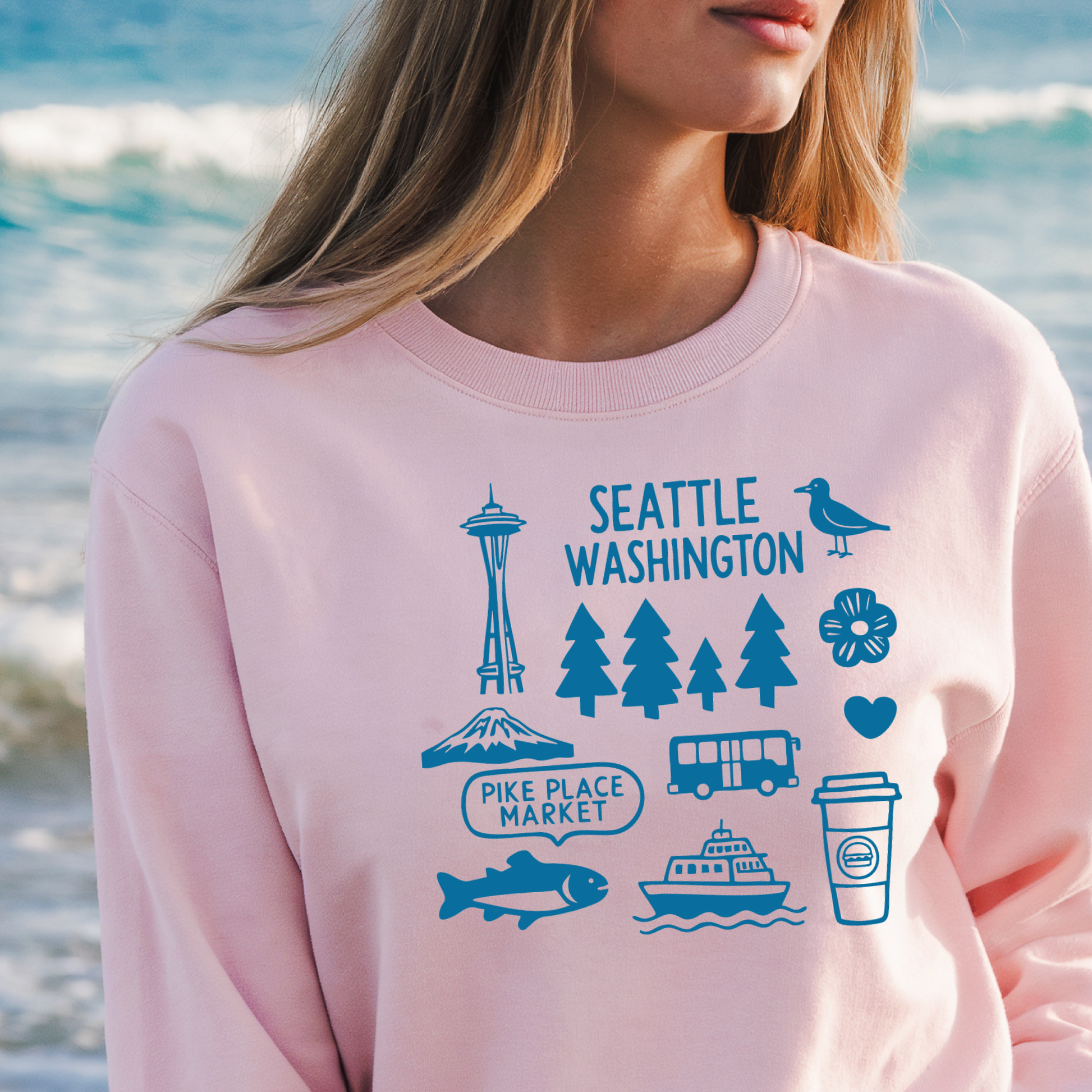Person wearing a pink sweatshirt with blue Seattle-themed graphics by the ocean.