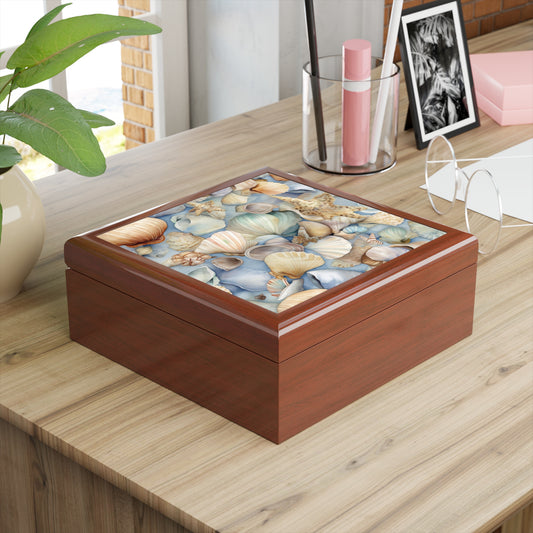 On a wooden desk sits the Coastal Shells Jewelry Box, a wooden piece with a colorful seashell lid. Nearby are eyeglasses, a potted plant, a photograph, and pens—a perfect nautical decor item or thoughtful bridesmaid gift.