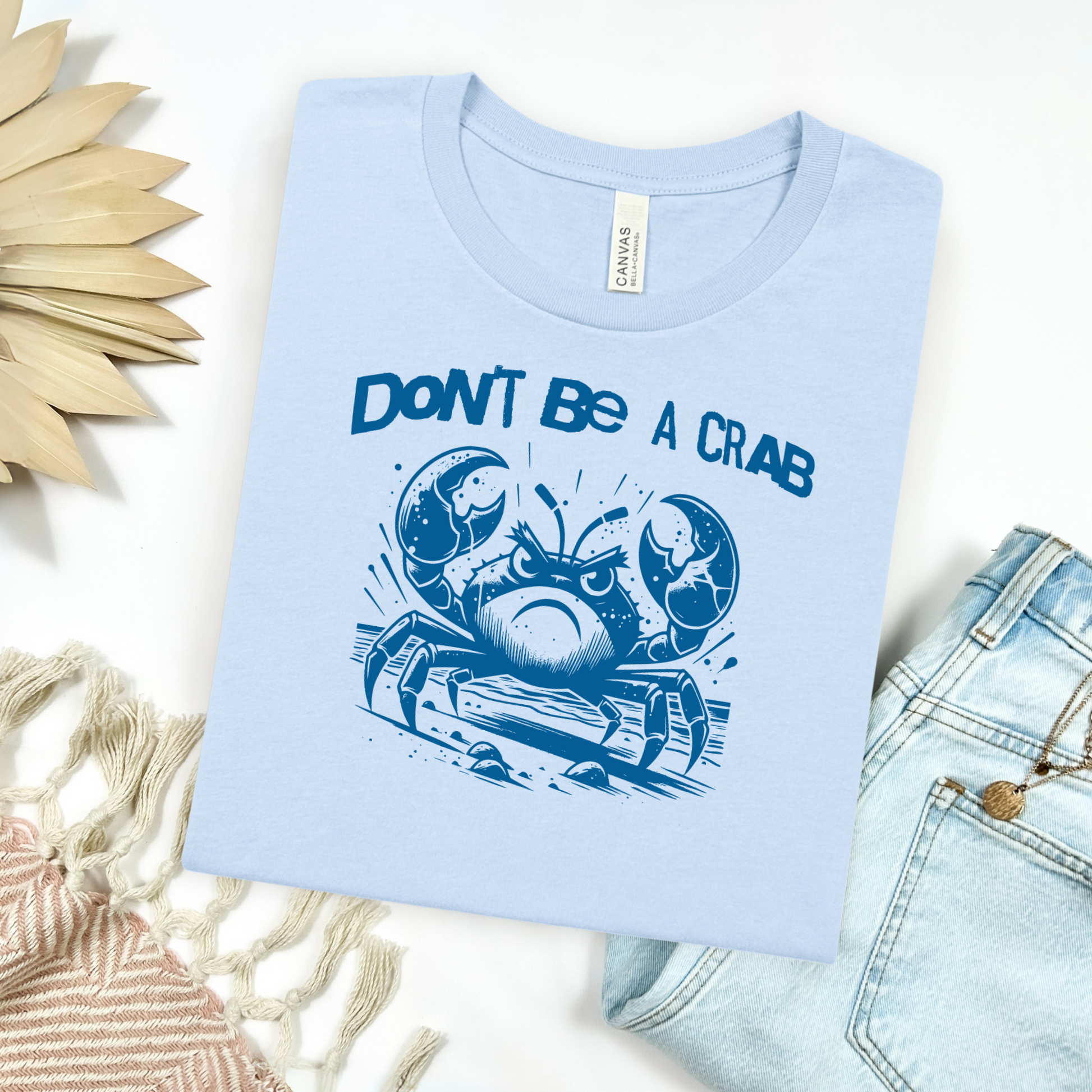 Bella Canvas 3001 "Don't Be A Crab" t shirt with cute angry crab in one color graphic. Picture on a flay lay mockup with pair of blue jeans and blanket and a wicker sun shaped accessory.