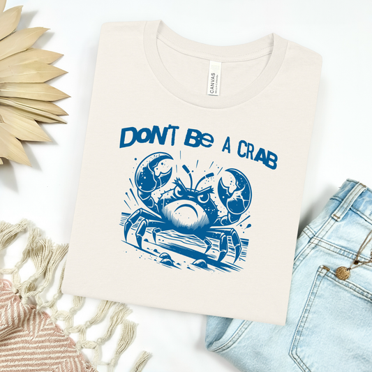 Bella Canvas 3001 "Don't Be A Crab" t shirt with cute angry crab in one color graphic. Picture on a flay lay mockup with pair of blue jeans and blanket and a wicker sun shaped accessory.