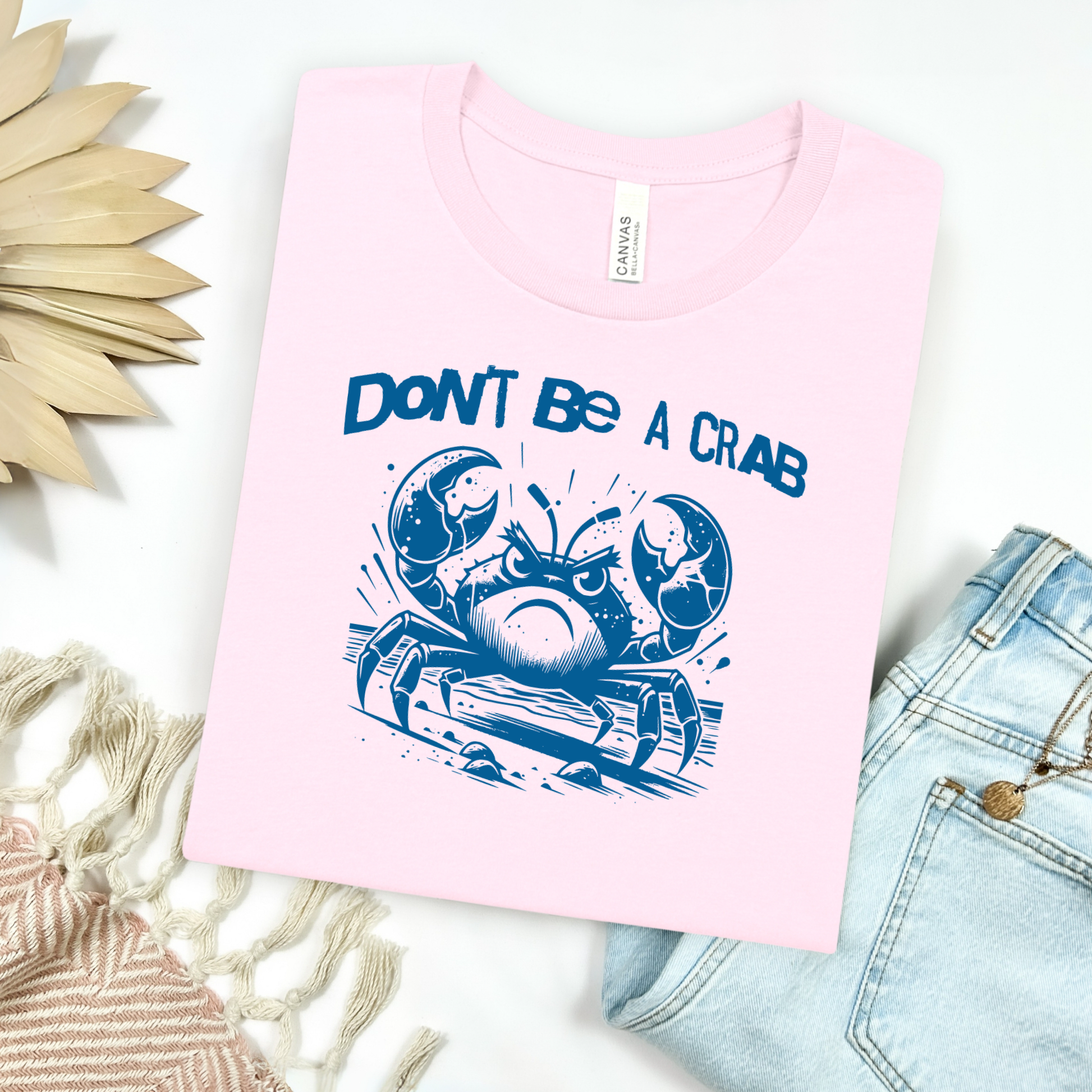 Bella Canvas 3001 "Don't Be A Crab" t shirt with cute angry crab in one color graphic. Picture on a flay lay mockup with pair of blue jeans and blanket and a wicker sun shaped accessory.