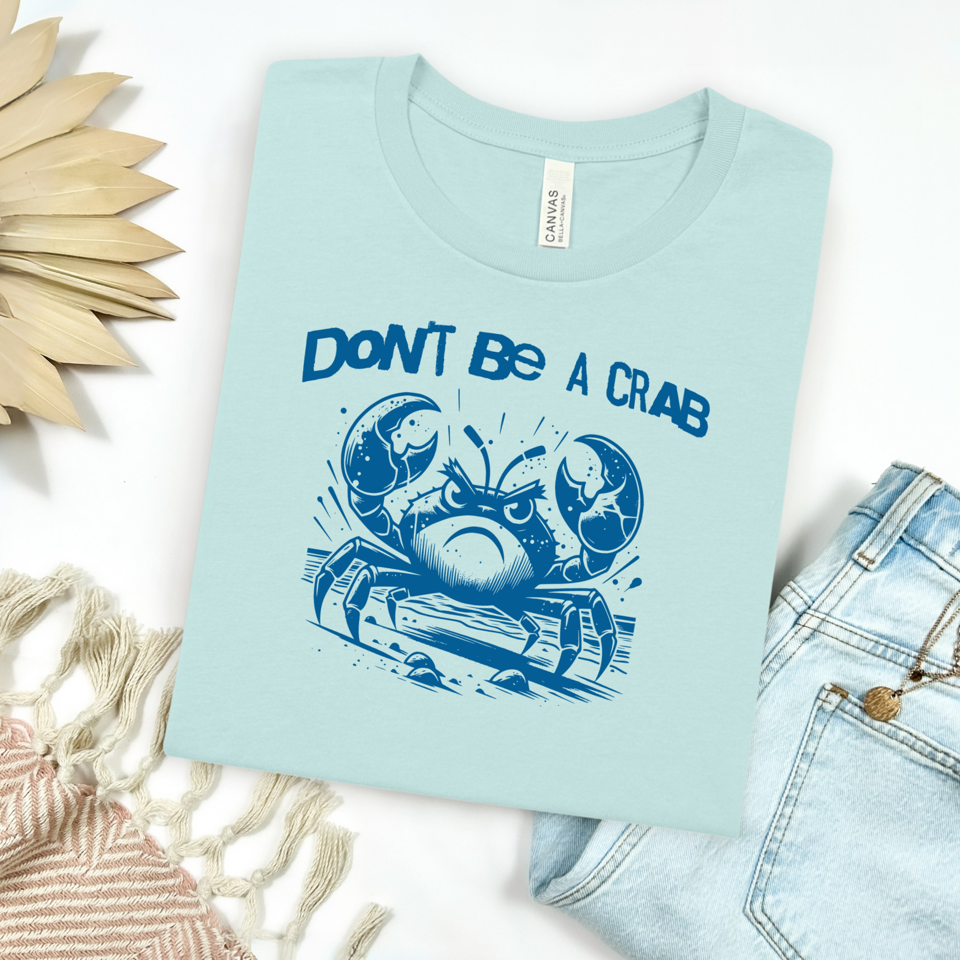 Bella Canvas 3001 "Don't Be A Crab" t shirt with cute angry crab in one color graphic. Picture on a flay lay mockup with pair of blue jeans and blanket and a wicker sun shaped accessory.