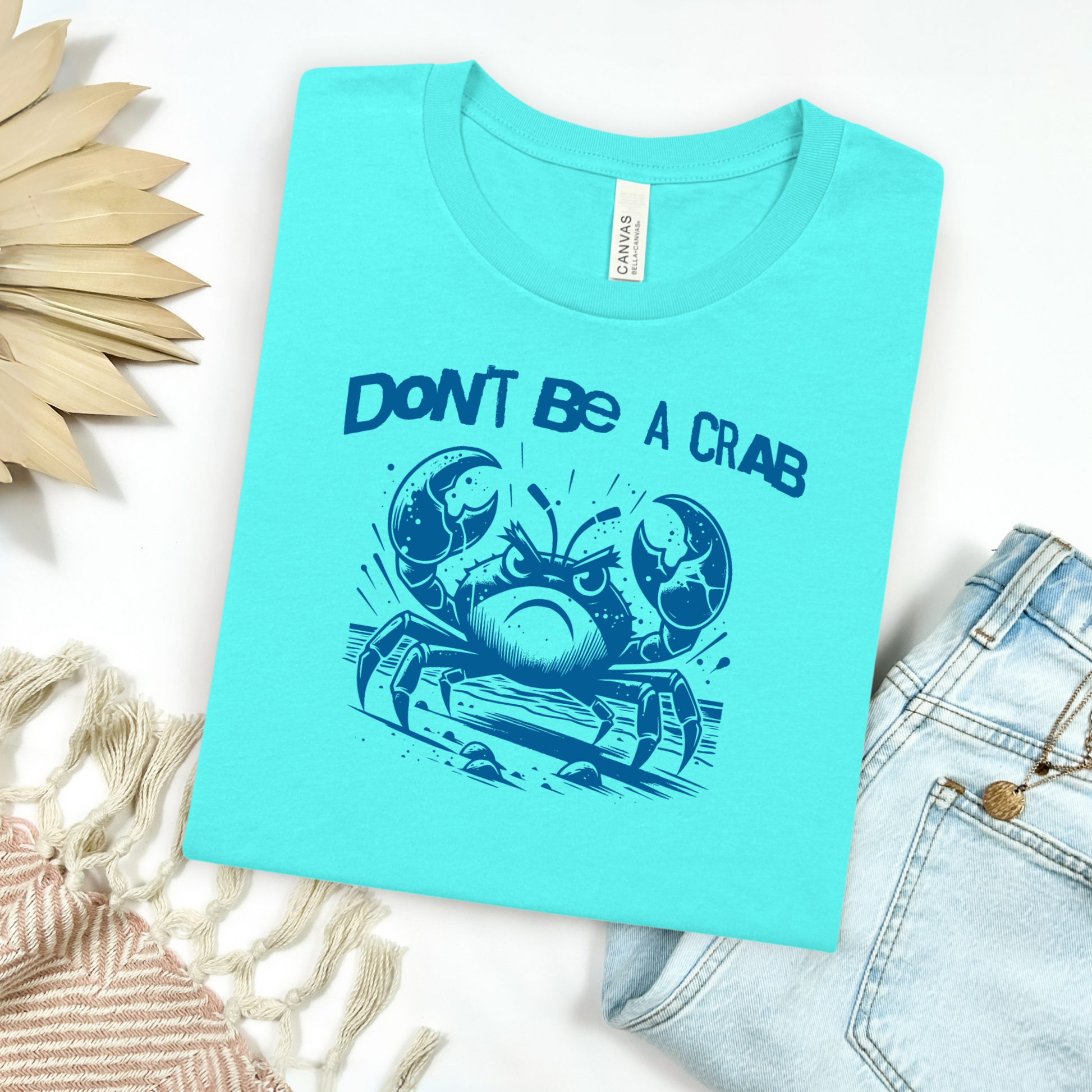 Bella Canvas 3001 "Don't Be A Crab" t shirt with cute angry crab in one color graphic. Picture on a flay lay mockup with pair of blue jeans and blanket and a wicker sun shaped accessory.