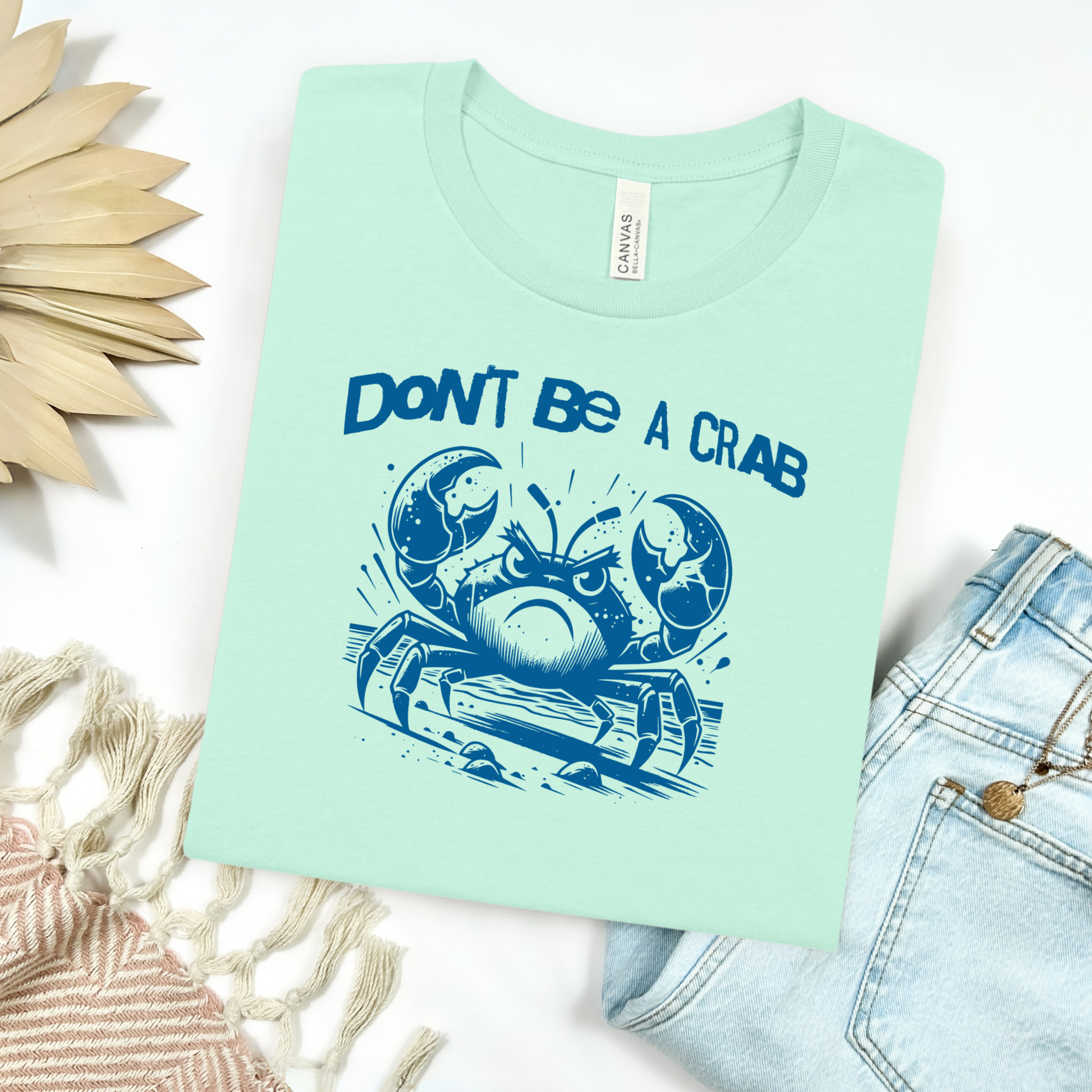 Bella Canvas 3001 "Don't Be A Crab" t shirt with cute angry crab in one color graphic. Picture on a flay lay mockup with pair of blue jeans and blanket and a wicker sun shaped accessory.