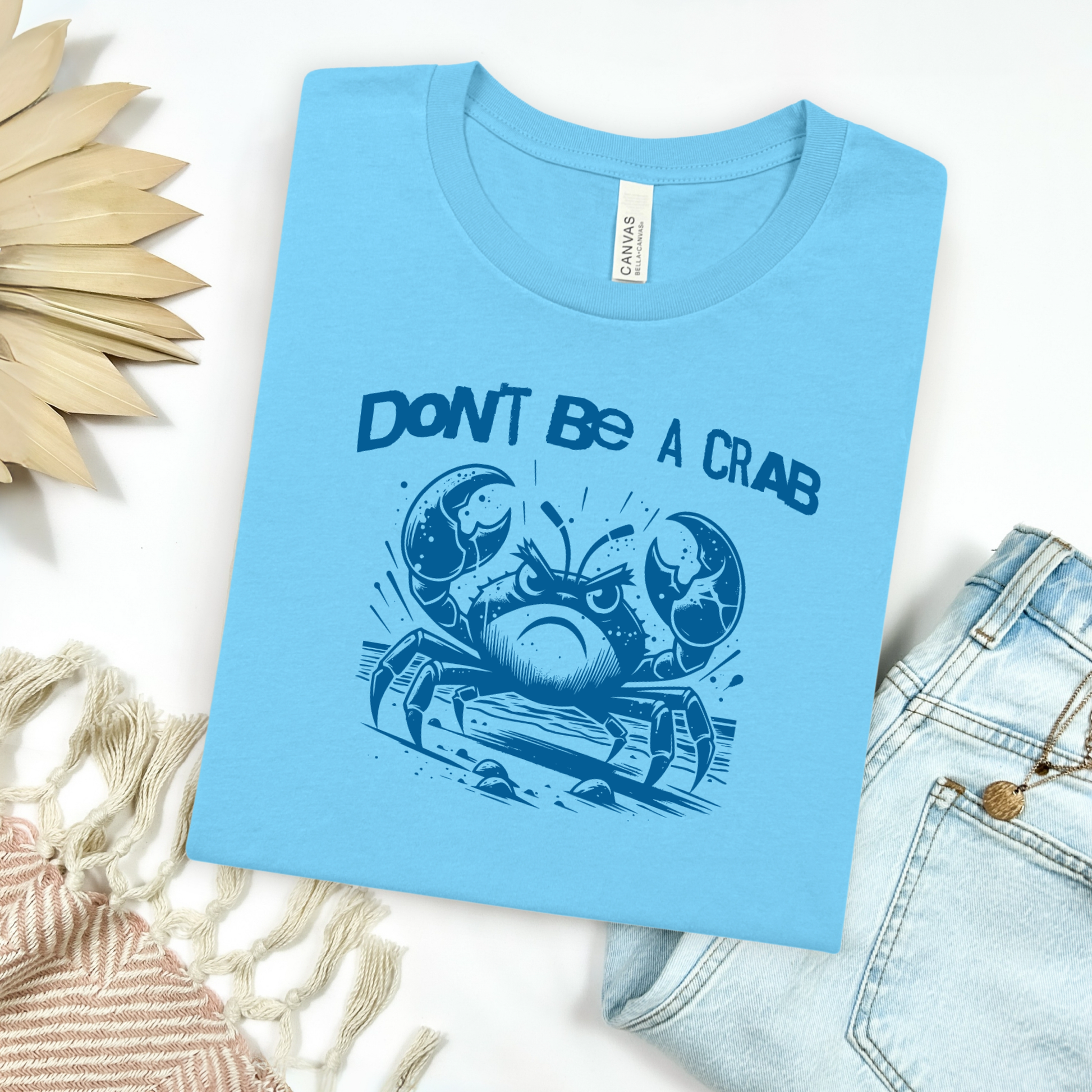 Bella Canvas 3001 "Don't Be A Crab" t shirt with cute angry crab in one color graphic. Picture on a flay lay mockup with pair of blue jeans and blanket and a wicker sun shaped accessory.