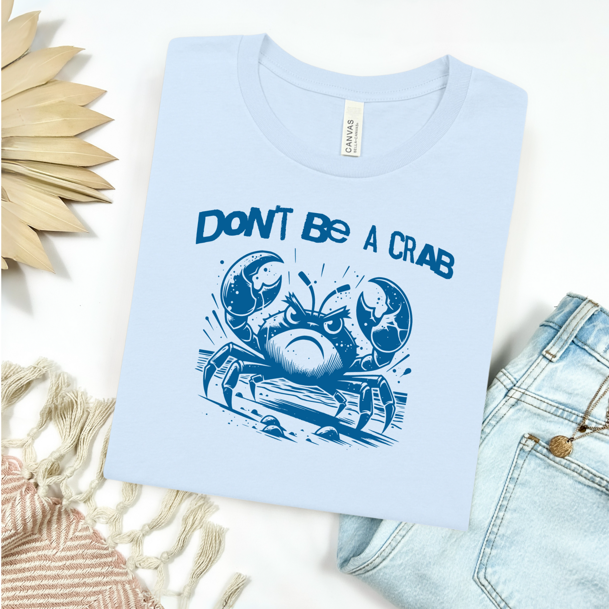 Bella Canvas 3001 "Don't Be A Crab" t shirt with cute angry crab in one color graphic. Picture on a flay lay mockup with pair of blue jeans and blanket and a wicker sun shaped accessory.