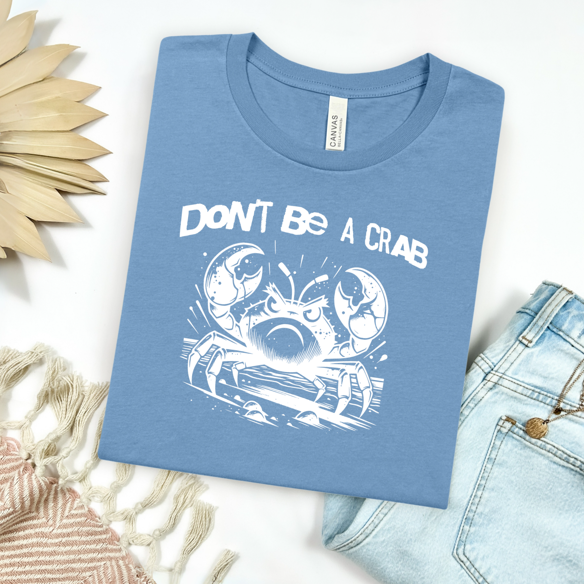 Bella Canvas 3001 "Don't Be A Crab" t shirt with cute angry crab in one color graphic. Picture on a flay lay mockup with pair of blue jeans and blanket and a wicker sun shaped accessory.