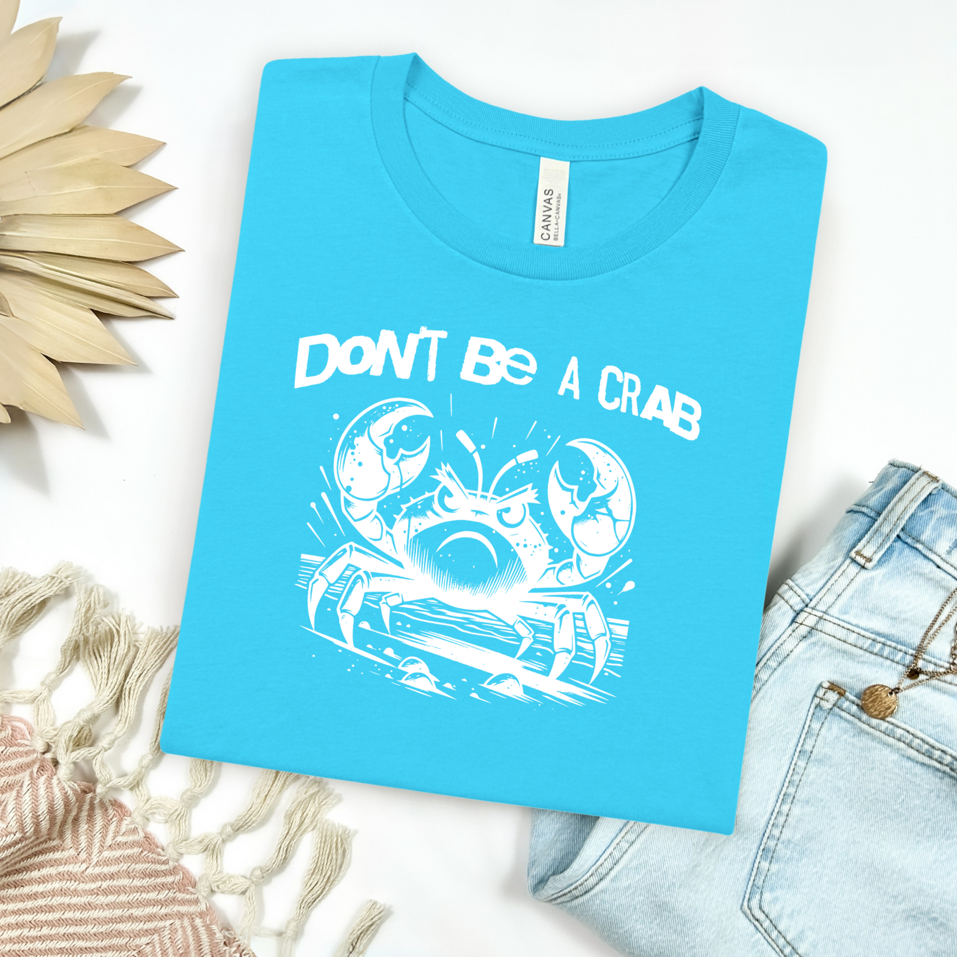 Bella Canvas 3001 "Don't Be A Crab" t shirt with cute angry crab in one color graphic. Picture on a flay lay mockup with pair of blue jeans and blanket and a wicker sun shaped accessory.