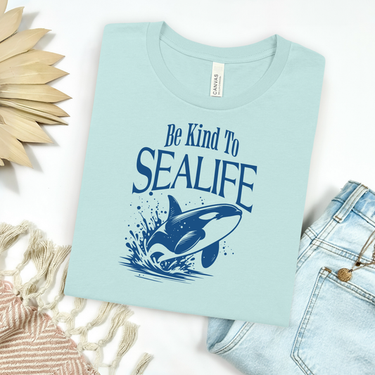 Bella + Canvas t-shirt with a one-color graphic of an orca whale jumping out of the ocean, displayed in a flat lay mockup alongside a pair of blue jeans, a cozy blanket, and a wicker sun-shaped accessory for a relaxed, beachy vibe.