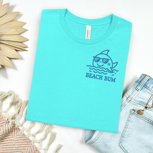 Bella and Canvas 3001 "Beach Bum" t shirt with dolphin and waves monoline one color graphic. Picture on a flay lay mockup with pair of blue jeans and blanket and a wicker sun shaped accessory.