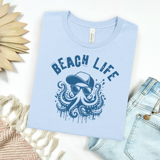 Bella Canvas 3001 " Beach Life" t shirt with a octopus wearing sunglasses and a baseball cap in a one color graphic. Picture on a flay lay mockup with pair of blue jeans and blanket and a wicker sun shaped accessory.