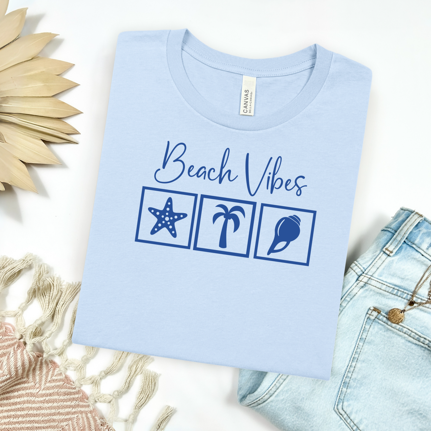 Bella and Canvas 3001 "Beach Vibes" t shirt with a starfish, palm tree, and a seashell graphics in monochrome color. Picture on a flay lay mockup with pair of blue jeans and blanket and a wicker sun shaped accessory.