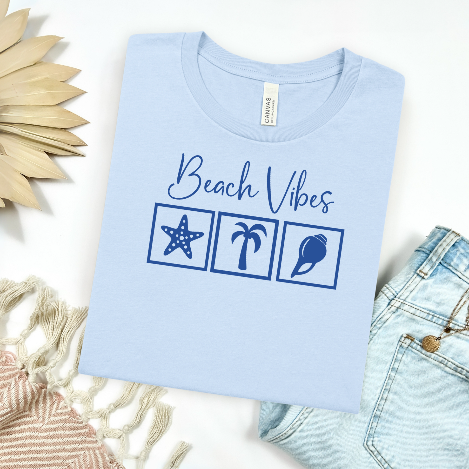 Bella and Canvas 3001 "Beach Vibes" t shirt with a starfish, palm tree, and a seashell graphics in monochrome color. Picture on a flay lay mockup with pair of blue jeans and blanket and a wicker sun shaped accessory.