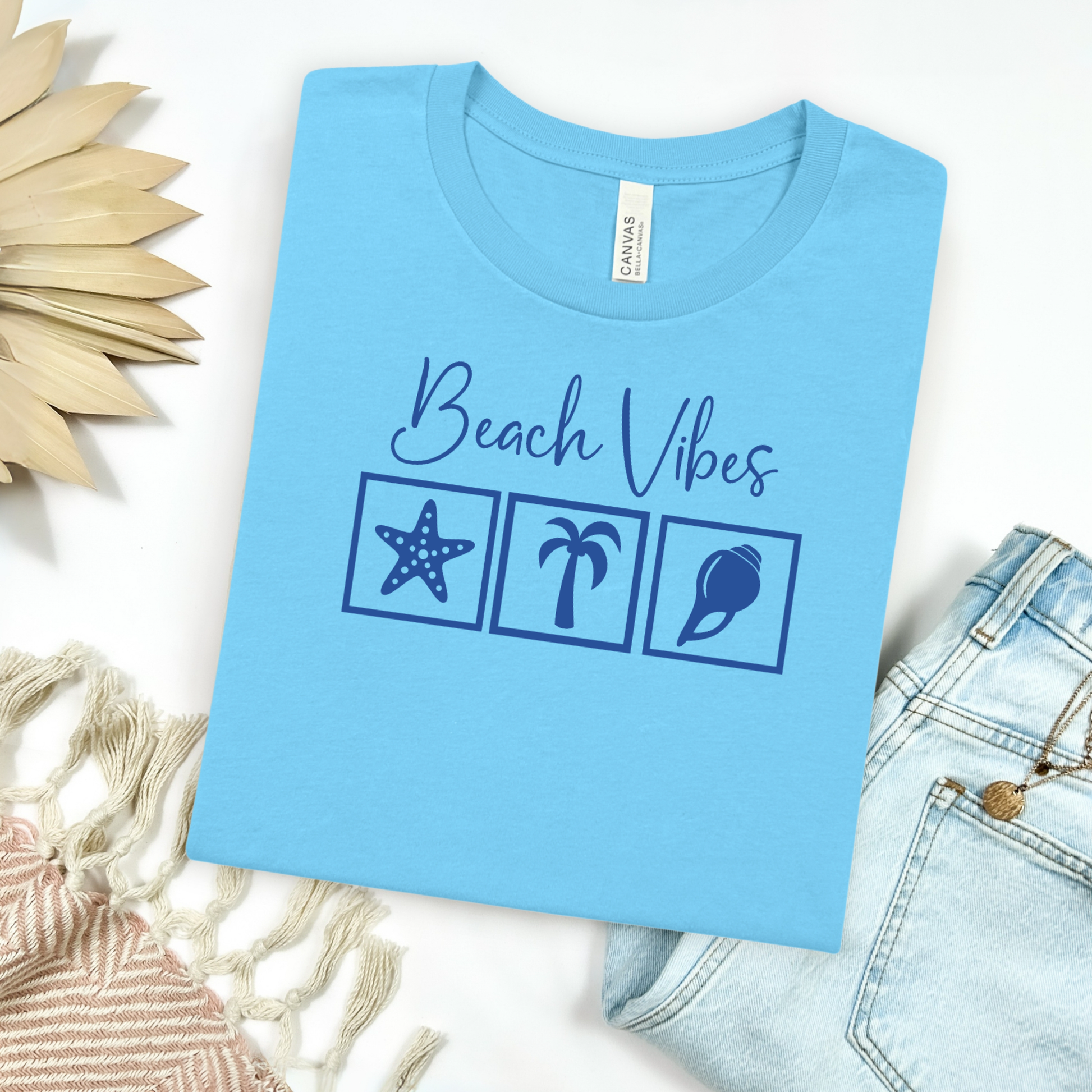 Bella and Canvas 3001 "Beach Vibes" t shirt with a starfish, palm tree, and a seashell graphics in monochrome color. Picture on a flay lay mockup with pair of blue jeans and blanket and a wicker sun shaped accessory.