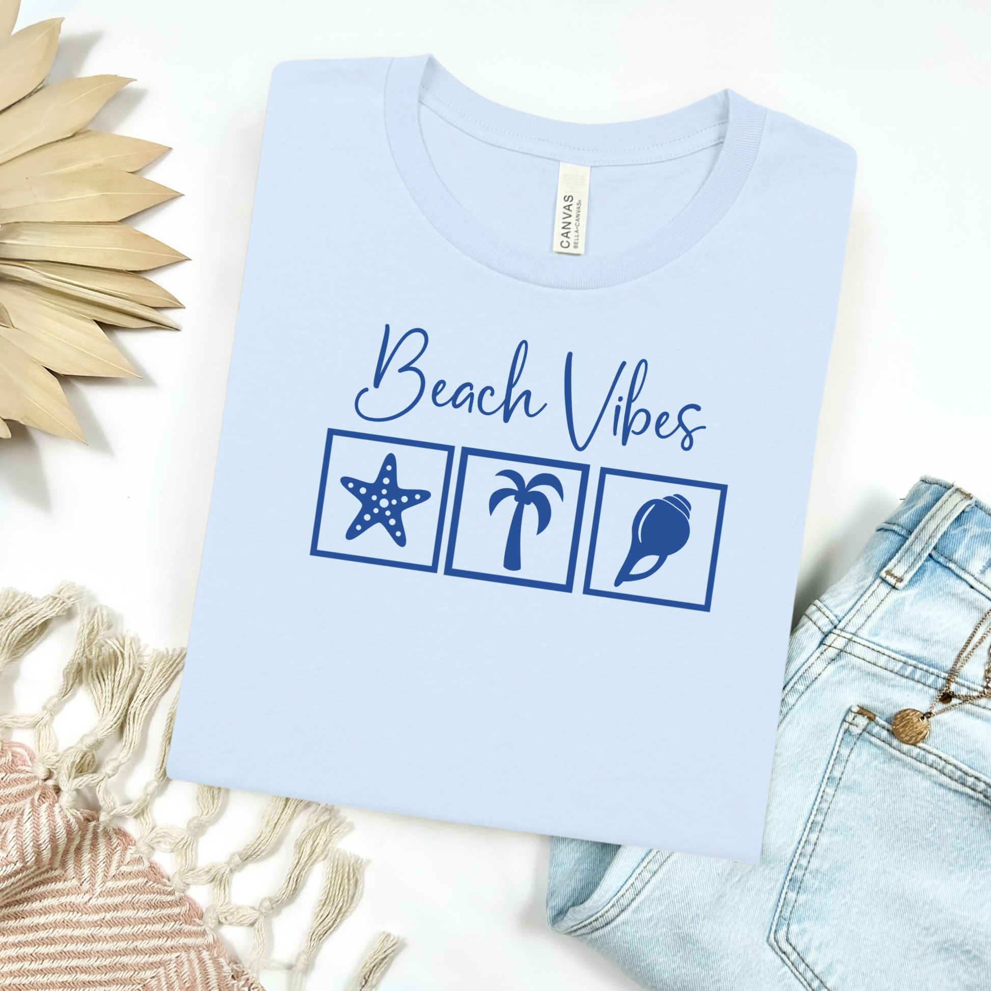 Bella and Canvas 3001 "Beach Vibes" t shirt with a starfish, palm tree, and a seashell graphics in monochrome color. Picture on a flay lay mockup with pair of blue jeans and blanket and a wicker sun shaped accessory.