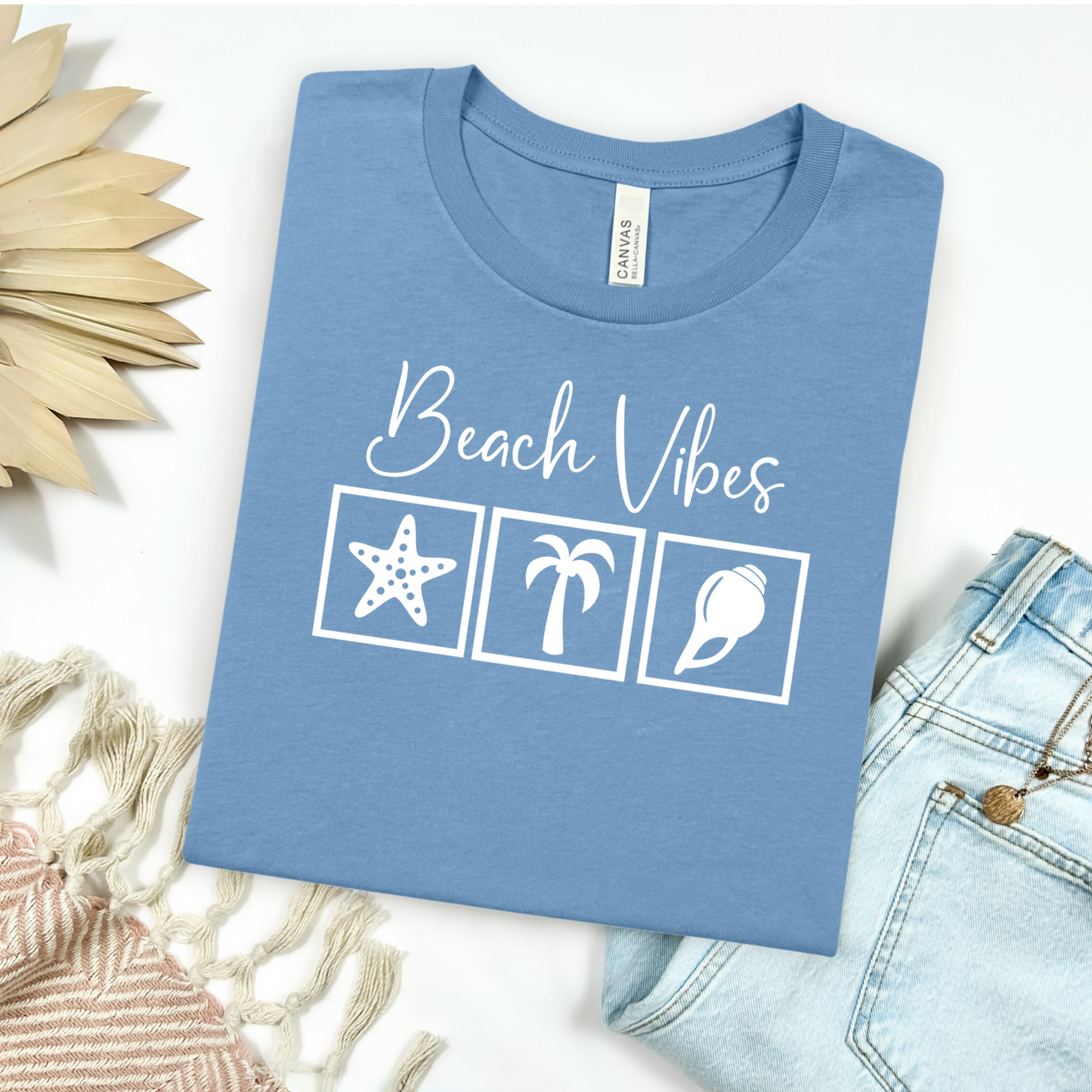 Bella and Canvas 3001 "Beach Vibes" t shirt with a starfish, palm tree, and a seashell graphics in monochrome color. Picture on a flay lay mockup with pair of blue jeans and blanket and a wicker sun shaped accessory.