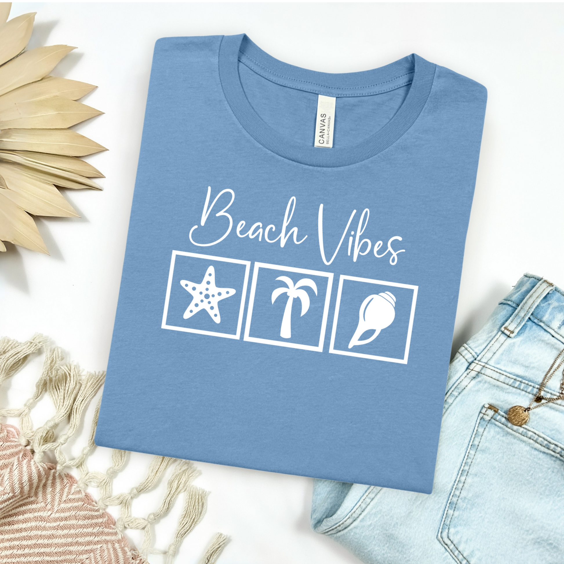 Bella and Canvas 3001 "Beach Vibes" t shirt with a starfish, palm tree, and a seashell graphics in monochrome color. Picture on a flay lay mockup with pair of blue jeans and blanket and a wicker sun shaped accessory.
