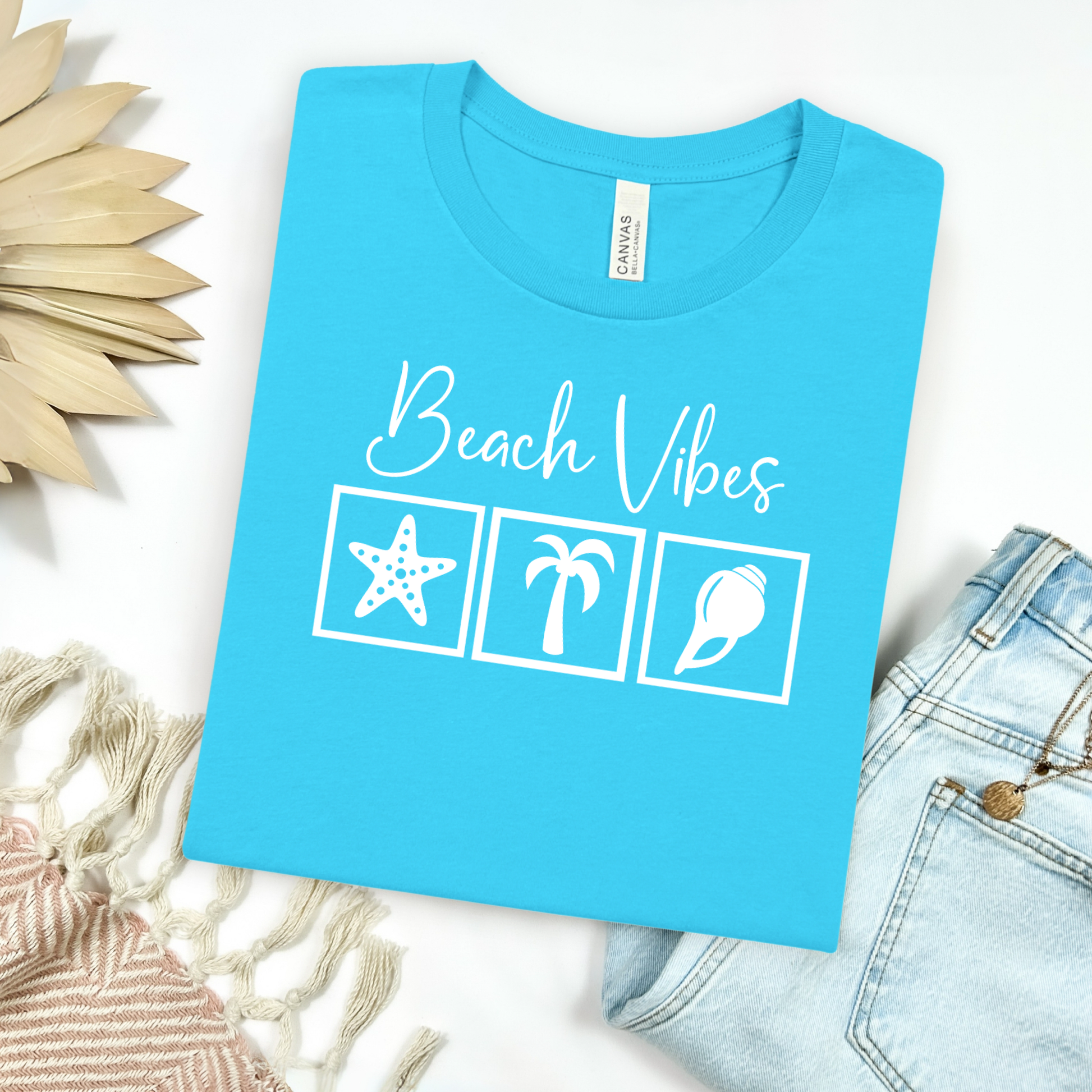 Bella and Canvas 3001 "Beach Vibes" t shirt with a starfish, palm tree, and a seashell graphics in monochrome color. Picture on a flay lay mockup with pair of blue jeans and blanket and a wicker sun shaped accessory.