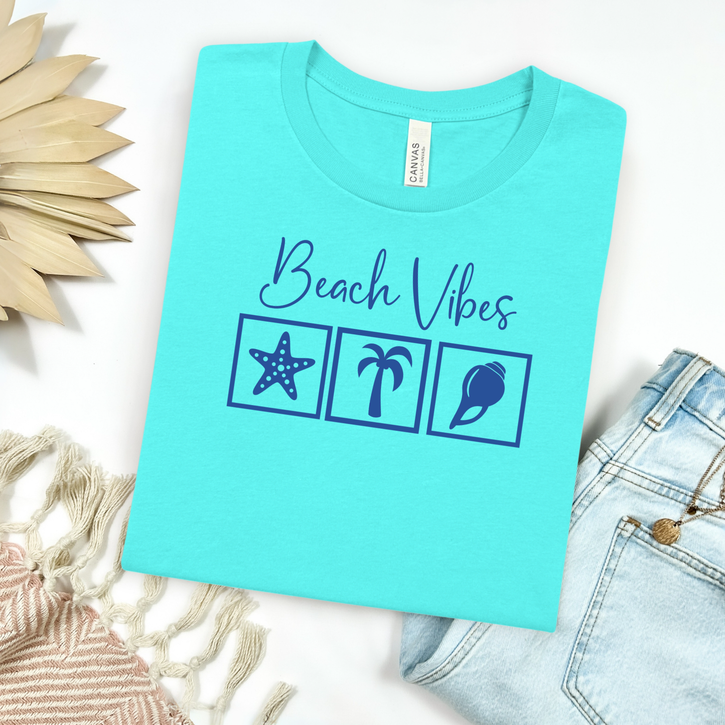 Bella and Canvas 3001 "Beach Vibes" t shirt with a starfish, palm tree, and a seashell graphics in monochrome color. Picture on a flay lay mockup with pair of blue jeans and blanket and a wicker sun shaped accessory.