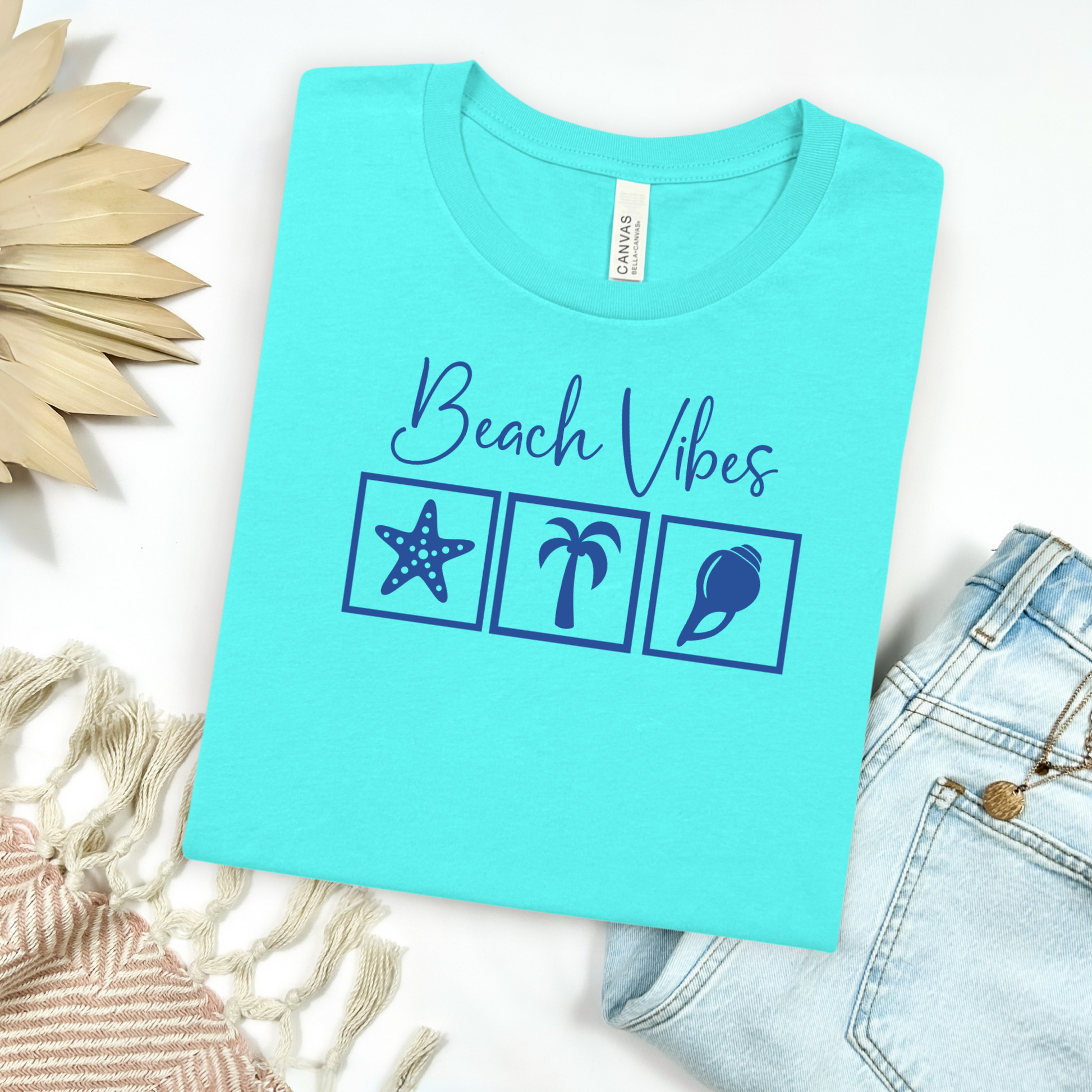 Bella and Canvas 3001 "Beach Vibes" t shirt with a starfish, palm tree, and a seashell graphics in monochrome color. Picture on a flay lay mockup with pair of blue jeans and blanket and a wicker sun shaped accessory.