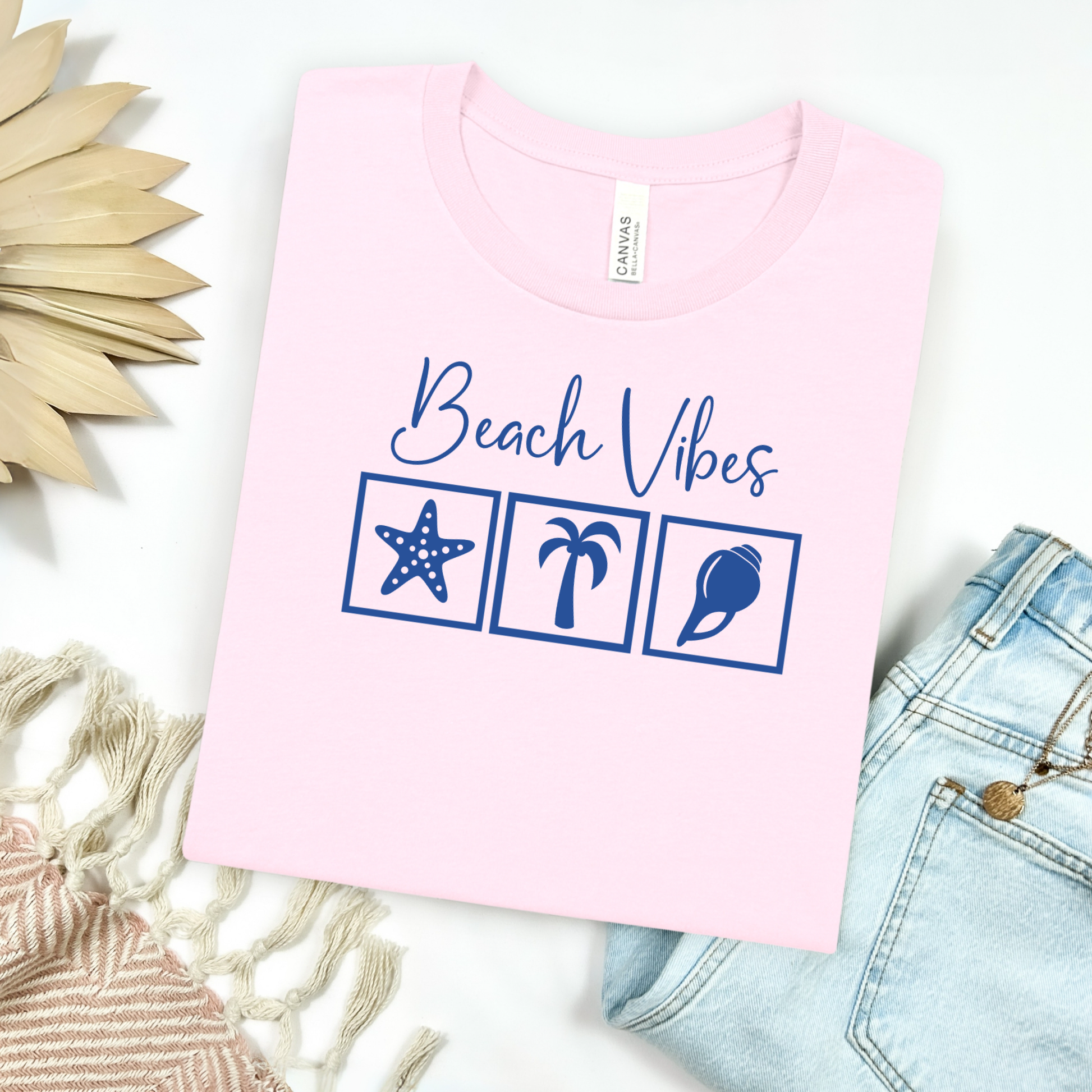 Bella and Canvas 3001 "Beach Vibes" t shirt with a starfish, palm tree, and a seashell graphics in monochrome color. Picture on a flay lay mockup with pair of blue jeans and blanket and a wicker sun shaped accessory.