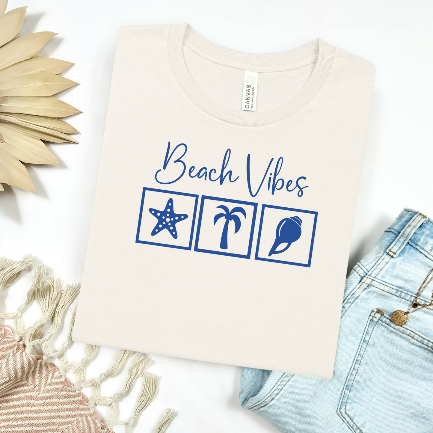 Bella and Canvas 3001 "Beach Vibes" t shirt with a starfish, palm tree, and a seashell graphics in monochrome color. Picture on a flay lay mockup with pair of blue jeans and blanket and a wicker sun shaped accessory.