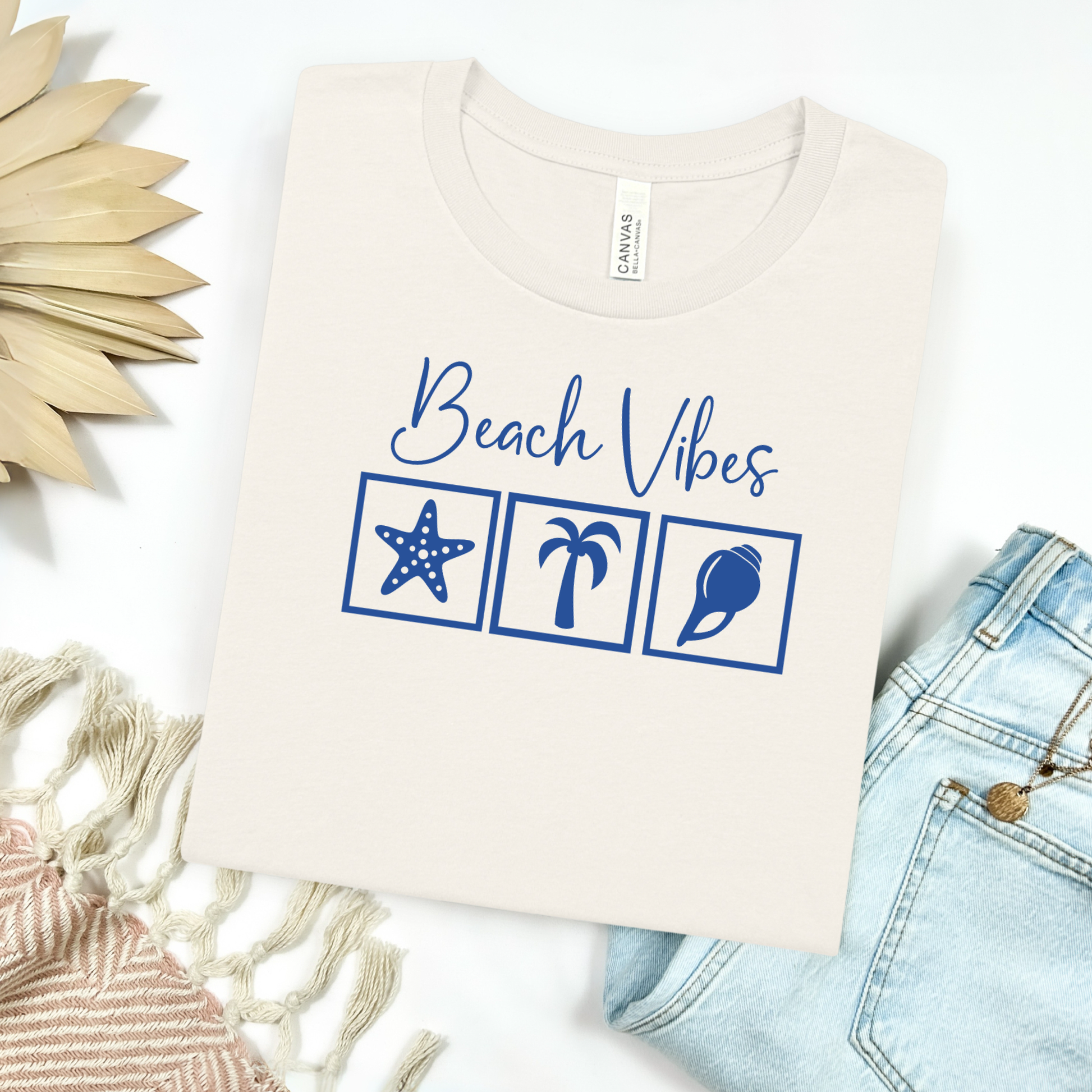 Bella and Canvas 3001 "Beach Vibes" t shirt with a starfish, palm tree, and a seashell graphics in monochrome color. Picture on a flay lay mockup with pair of blue jeans and blanket and a wicker sun shaped accessory.