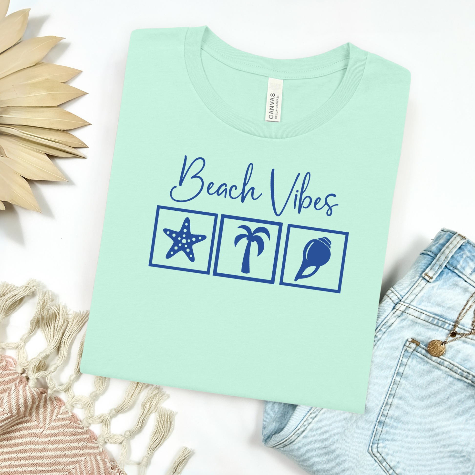 Bella and Canvas 3001 "Beach Vibes" t shirt with a starfish, palm tree, and a seashell graphics in monochrome color. Picture on a flay lay mockup with pair of blue jeans and blanket and a wicker sun shaped accessory.